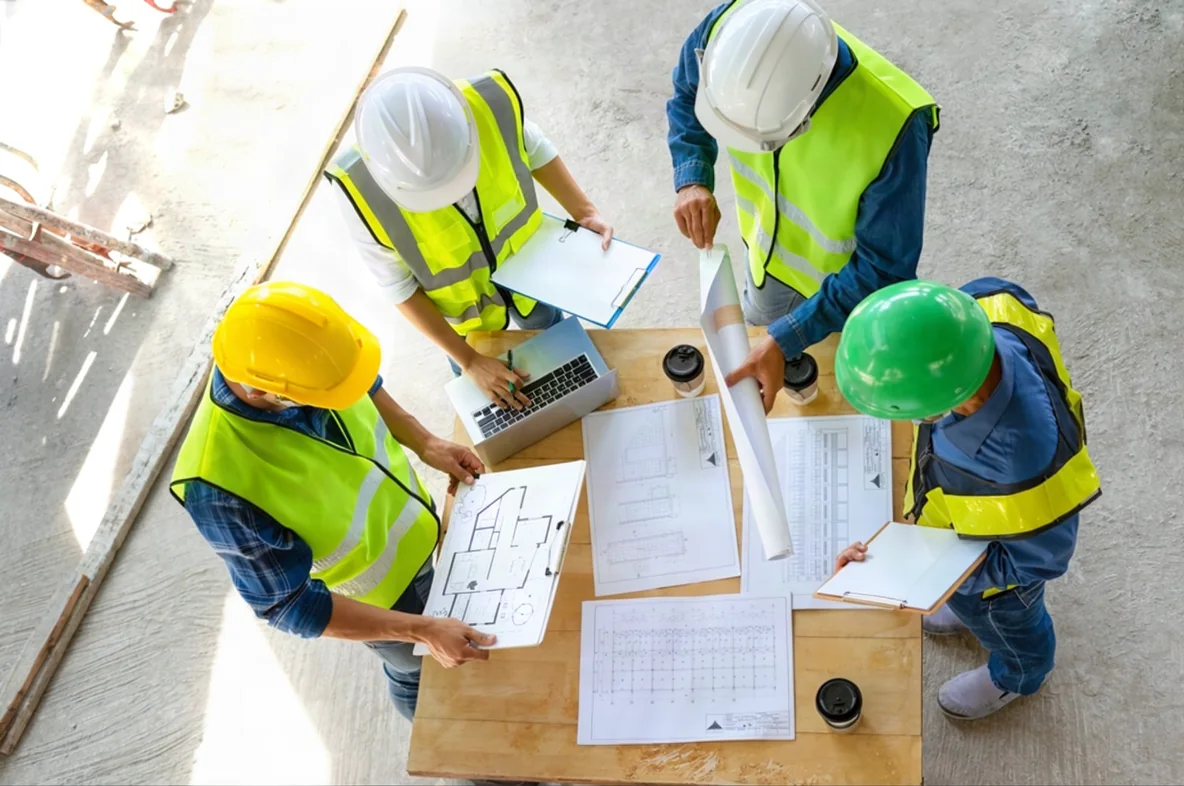 Construction professionals gathered around drawings and a laptop at a jobsite planning meeting