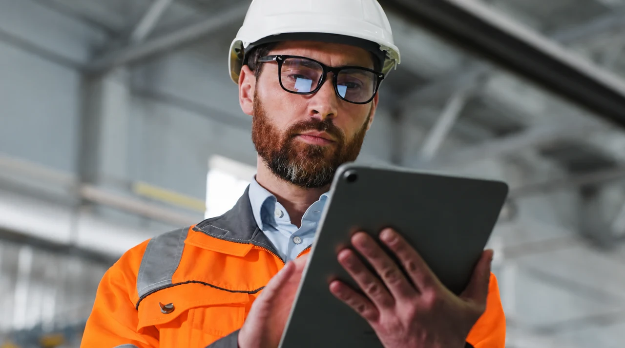 Construction manager wearing an orange safety vest and white hard hat, reviewing a tablet inside an unfinished building